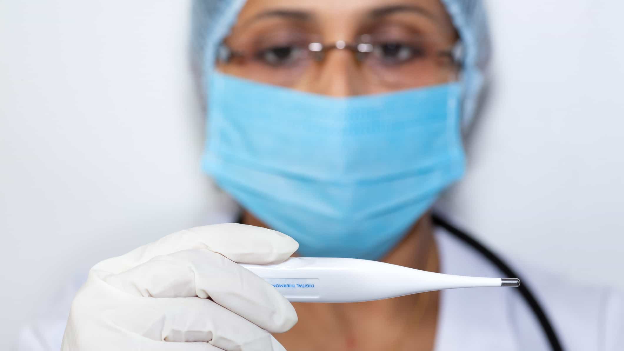 a medical person in protective clothing, rubber gloves, a mask and hair covering holds a digital thermometer up to the front of the picture as if to take someone's temperature.