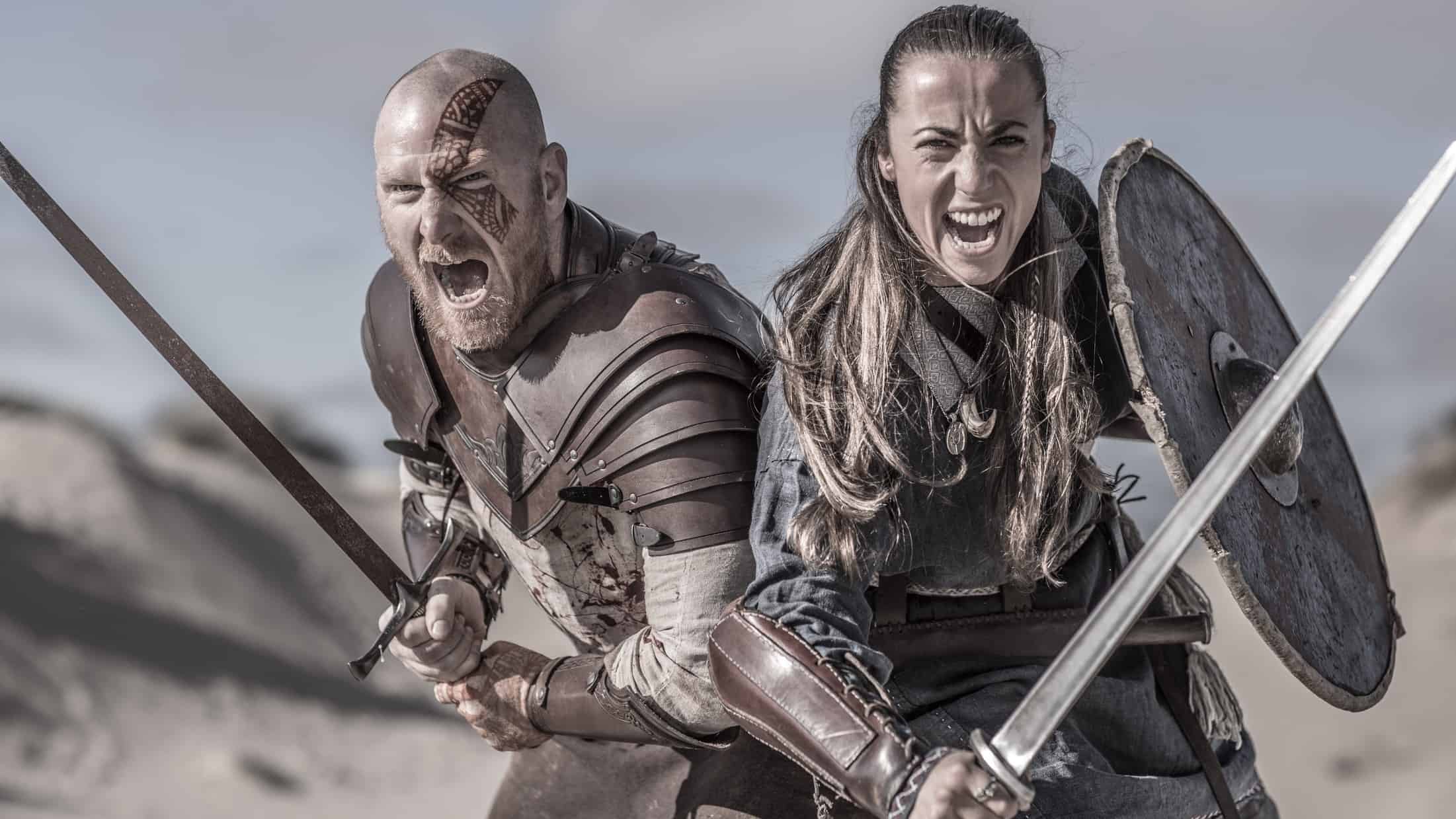 two medieval style warriors wearing armour and carrying shields and swords stand side by side as if joined at the hip with fericious, wide mouthed expressions on their faces as if ready for battle.