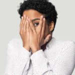 a woman with her hands over her face splits her fingers over one eye so she can peep through them.