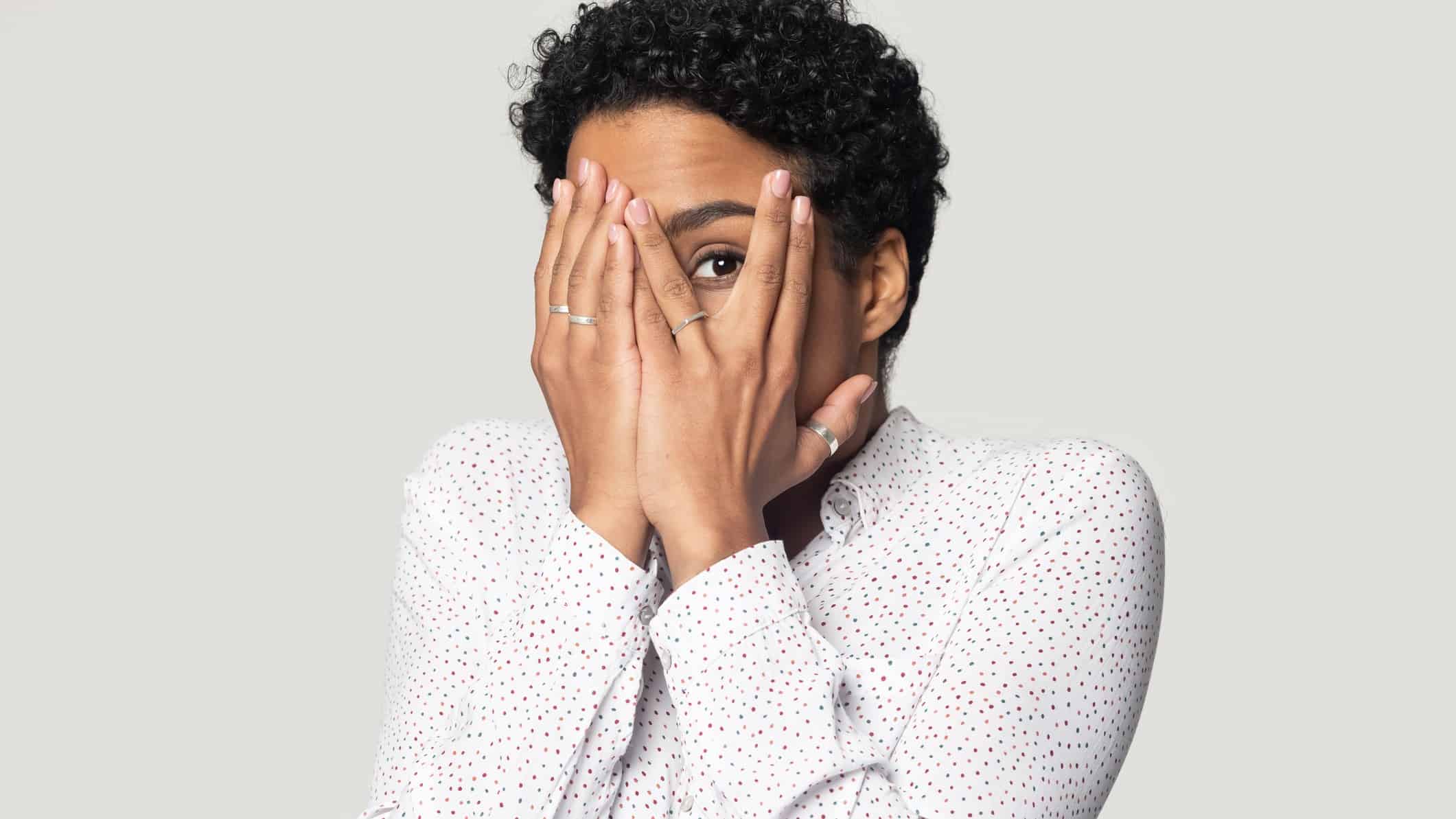 a woman with her hands over her face splits her fingers over one eye so she can peep through them.