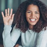 a woman sits at her desk with her hand up as if saying 'pick me' as she smiles widely.