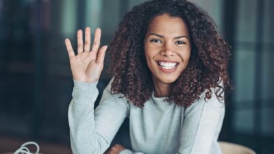 a woman sits at her desk with her hand up as if saying 'pick me' as she smiles widely.