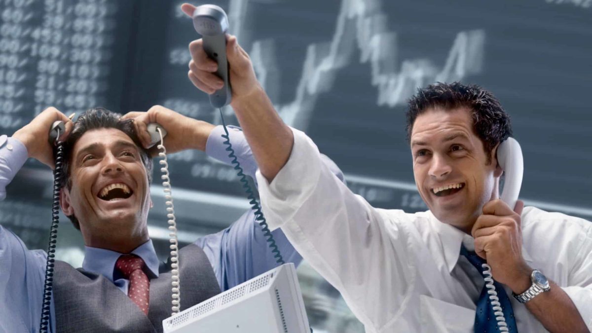 Two men look excited on the trading floor as they hold telephones to their ears and one points upwards.