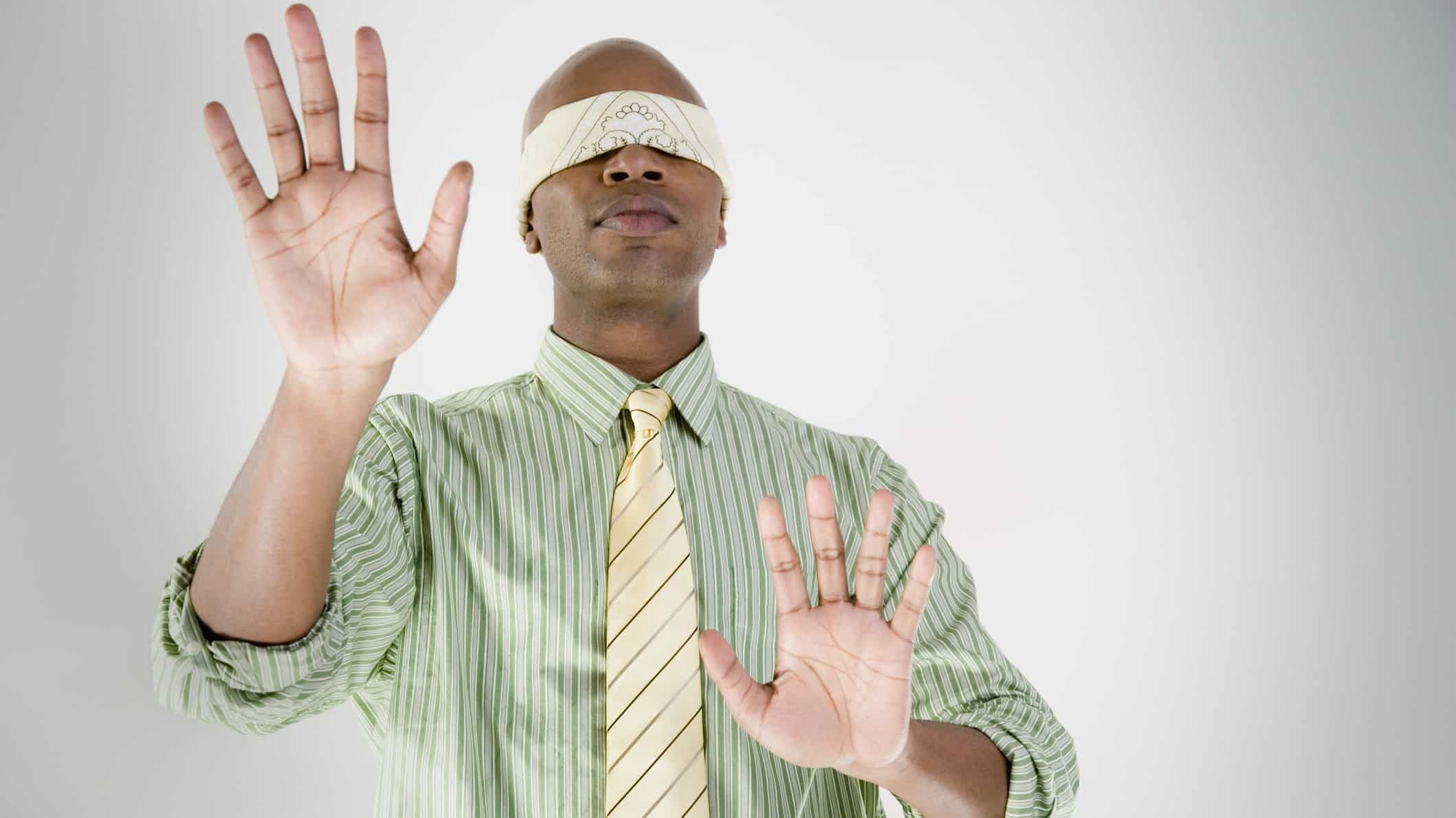 a man wearing a blindfold and business shirt and tie holds his hands up as if to feel his way.