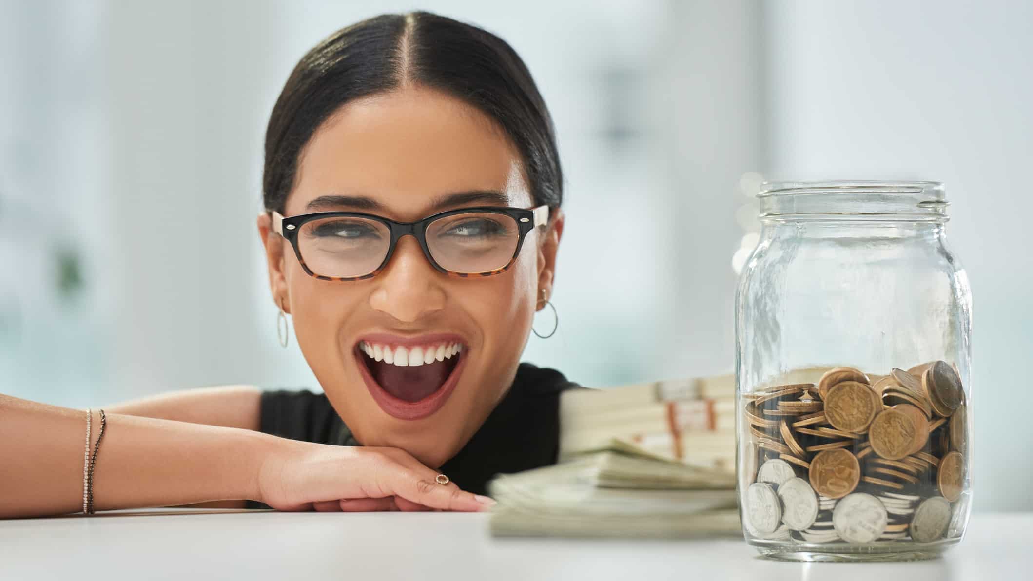 a woman with a huge happy smile on her face eyes a jar of coins next to her on a table.