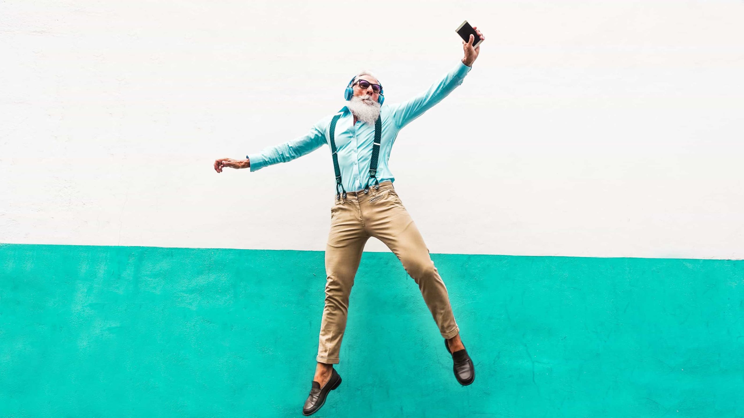 A cool older man leaps in the air wearing headphones and holding his mobile phone.