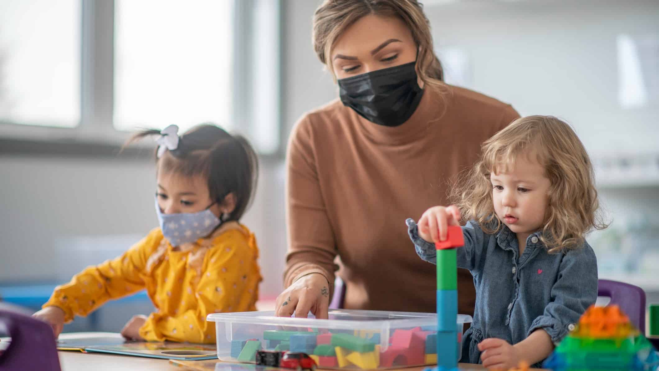 a childcare teacher wearing a face mask sits at a table with two small children playing with blocks. One of the children is also wearing a facemask.
