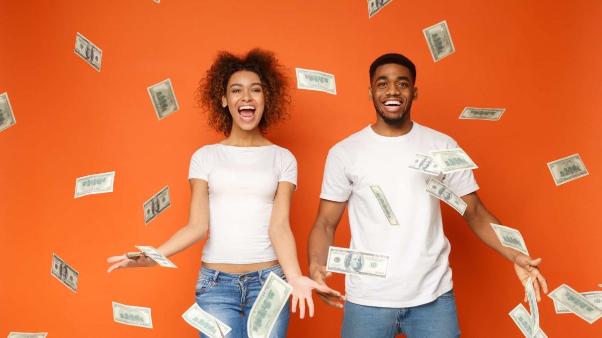 Happy young man and woman throwing dividend cash into air in front of orange background.