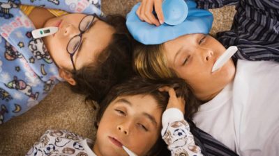three children lie on the floor with heads together with thermometers in their mouths. They are looking sick with eyes half closed and one is holding a cold pack to his forehead.