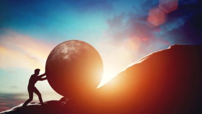 Man pushing large rock up hill with sunrise in background