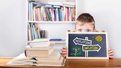 Young boy sitting at desk holding chalkboard sign with 'Deflation' and 'Inflation' written on it.