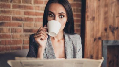 Female ASX travel shares investor with surprised expression drinks a cup of tea while reading the newspaper at her desk