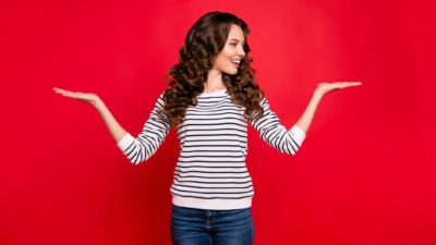 Woman in striped long sleeved top holds both hands up and looks to one side signifying a comparison between two ASX shares