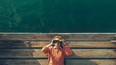 Woman in pink sweater lying on dock with binoculars to her eyes