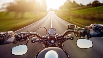 Photo from motorcycle rider's perspective looking at handlebars and road with green fields either side