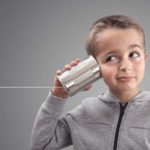 A young boy in a grey zip-up jumper has a tin can connected to a string pressed to his ear.
