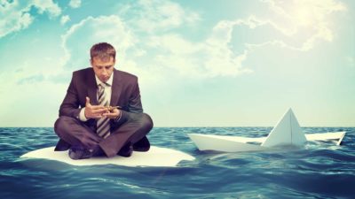 Man in business suit sits on sinking raft while looking at phone