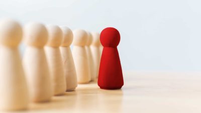 small red wooden peg doll standing ahead of group of neutral coloured peg dolls