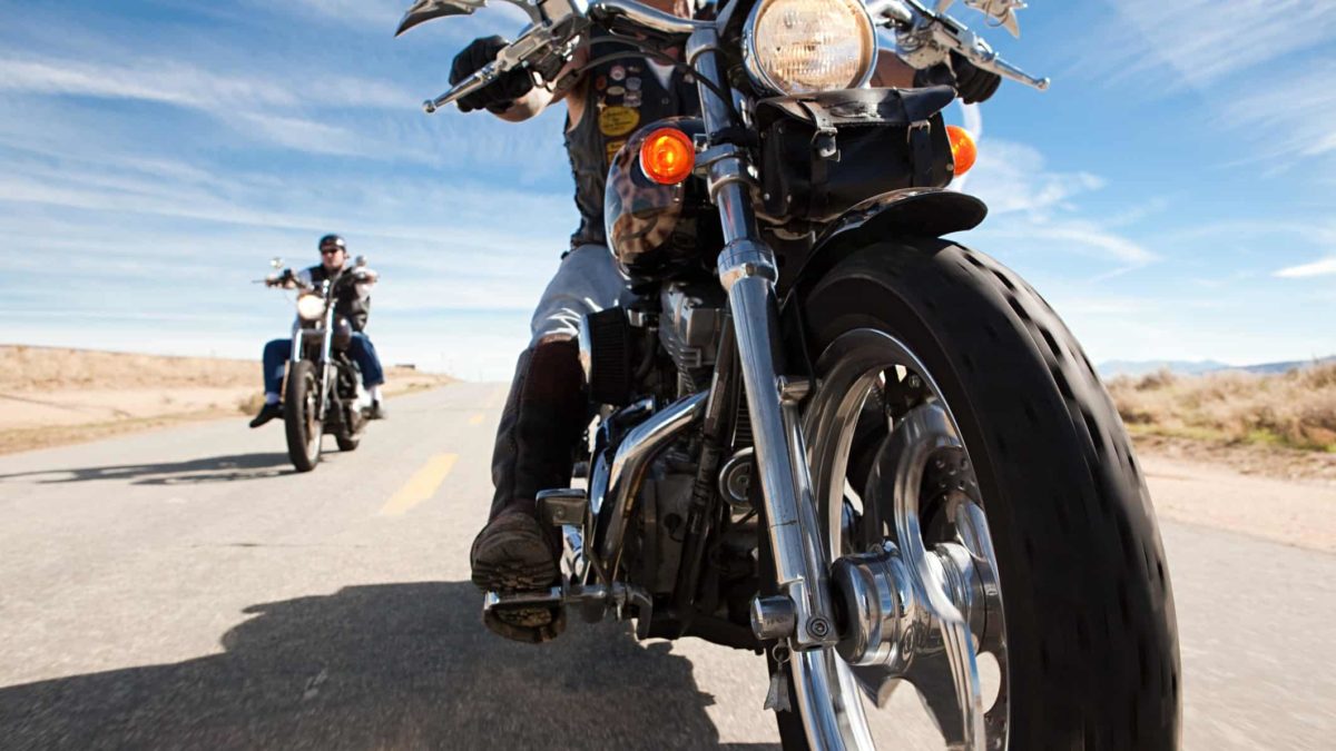 a close up of a motorcycle's front wheel and body on the open road with another motorcycle rider in the background cruising behind the leading driver.