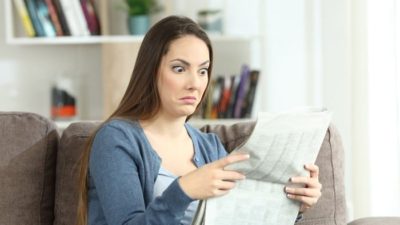 Woman sitting on couch holding newspaper with shocked expression on face