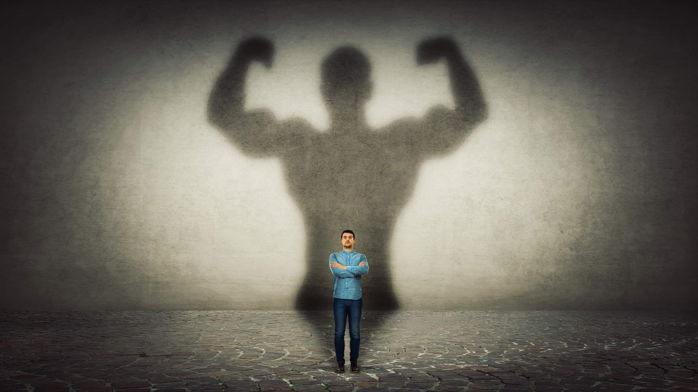 A man stands with arms crossed in front of a giant shadow of a body builder representing ASX small-cap stocks.