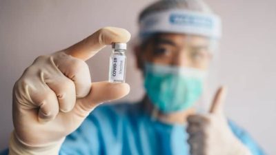 doctor making thumbs up gesture and holding vial labelled 'covid-19 vaccine' representing covid shares