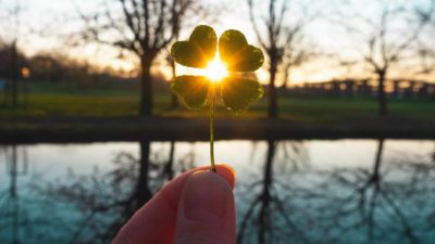 hand holding four leaf clover against backdrop of sun setting over lake