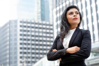 A businesswoman stands with arms crossed in a powerful pose with the city buildings behind her.