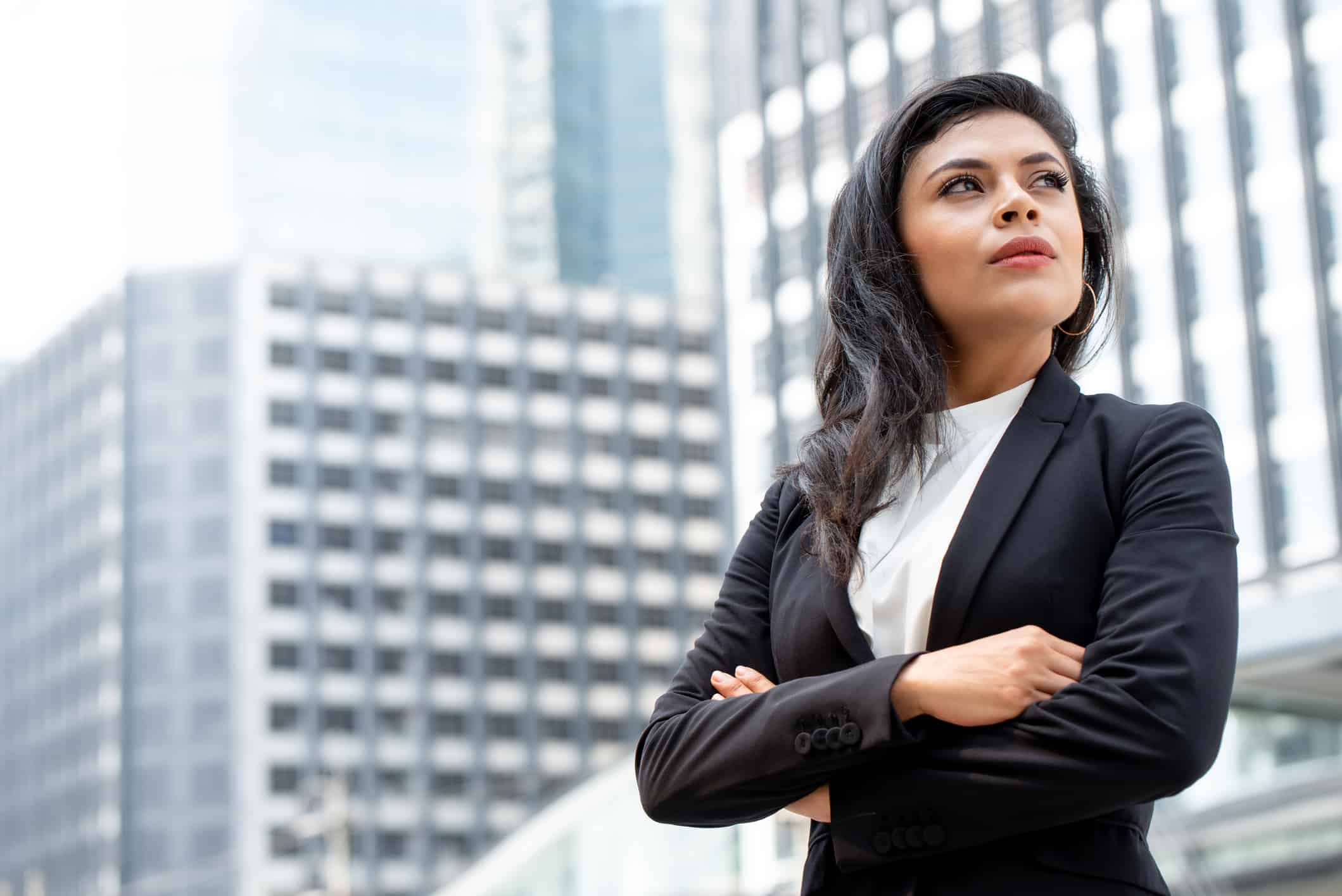 A businesswoman stands with arms crossed in a powerful pose with the city buildings behind her.