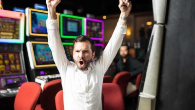 Man sitting at poker machine celebrates a win by raising his arms straight up in the air.