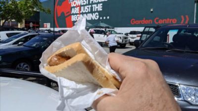 Bunnings stock represented by hand holding a sausage in bread against backdrop of Bunnings store