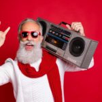 A cool older dude with a big white beard and wearing a red scarf holds a boombox stereo on his shoulder and makes rock'n'roll devil fingers with his other hand.