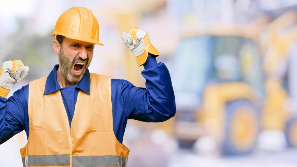 A mining worker wearing a hard hat, orange high vis vest and blue long-sleeved shirt raises his fists in celebration with an excited expression on his face