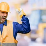 A mining worker wearing a hard hat, orange high vis vest and blue long-sleeved shirt raises his fists in celebration with an excited expression on his face