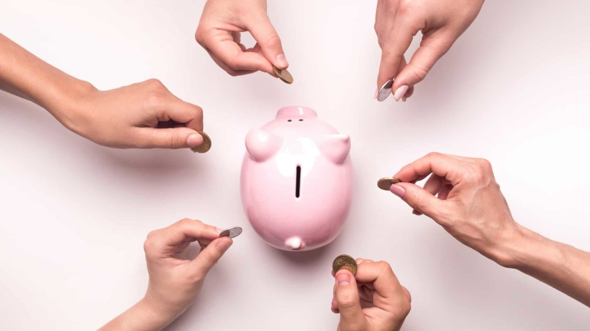 A piggy bank is surround by hands preparing to pay coins into the slot, representing a company capital raisingh in asx share price represented by multiple hands all placing coins in a piggy bank