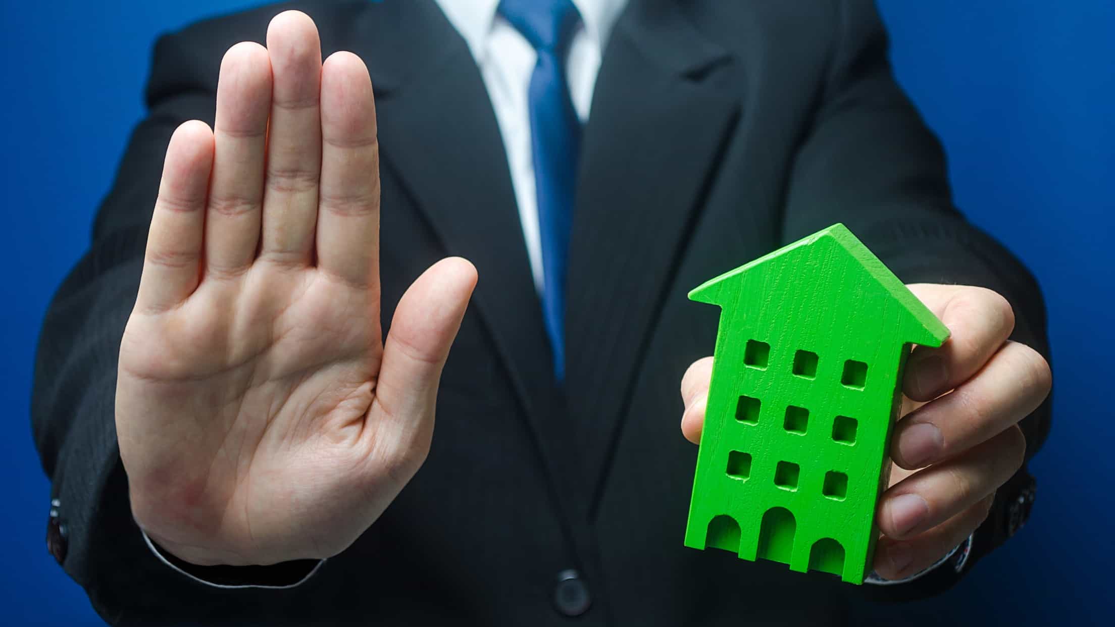real estate investment trust trading halt represented by man holding hand up in stop motion and holding wooden block in the shape of a house