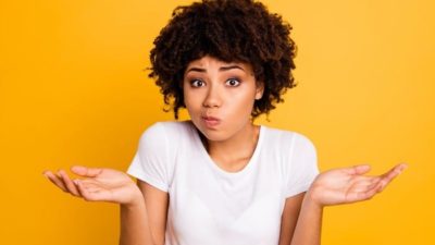 A woman with black afro hair and wearing a white t-shirt shrugs and purses her lips