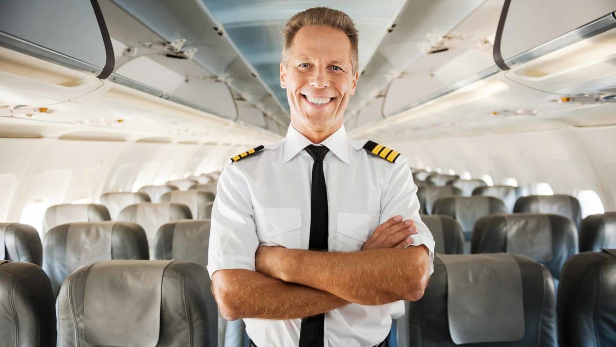 A pilot stands in an empty passenger cabin smiling with his arms crossed looking excited