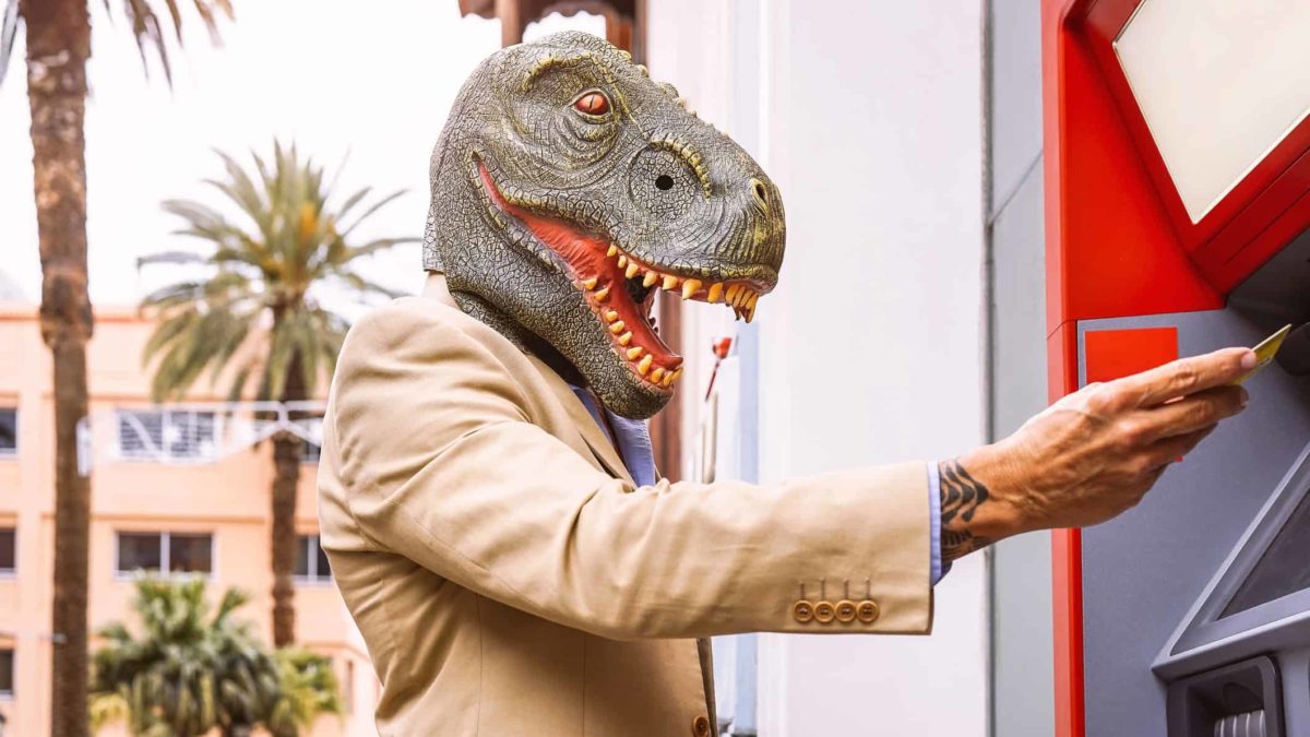 asx shares versus cash represented by man in dinosaur mask withdrawing cash from atm