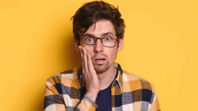 A man wearing glasses and a checkered shirt looks gobsmacked as he puts his hand to his cheek, representing the fall of the Zip share price is cheek