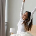 A woman wakes up after sleeping soundly, stretching her arms high sitting in bed.
