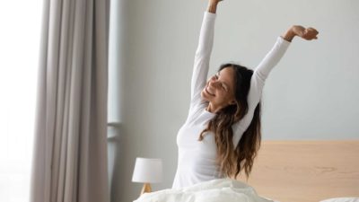 A woman wakes up after sleeping soundly, stretching her arms high sitting in bed.
