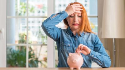 woman looking scared as she cradle a piggy bank and adds a coin, indictating a share investor holding on amid a volatile ASX market