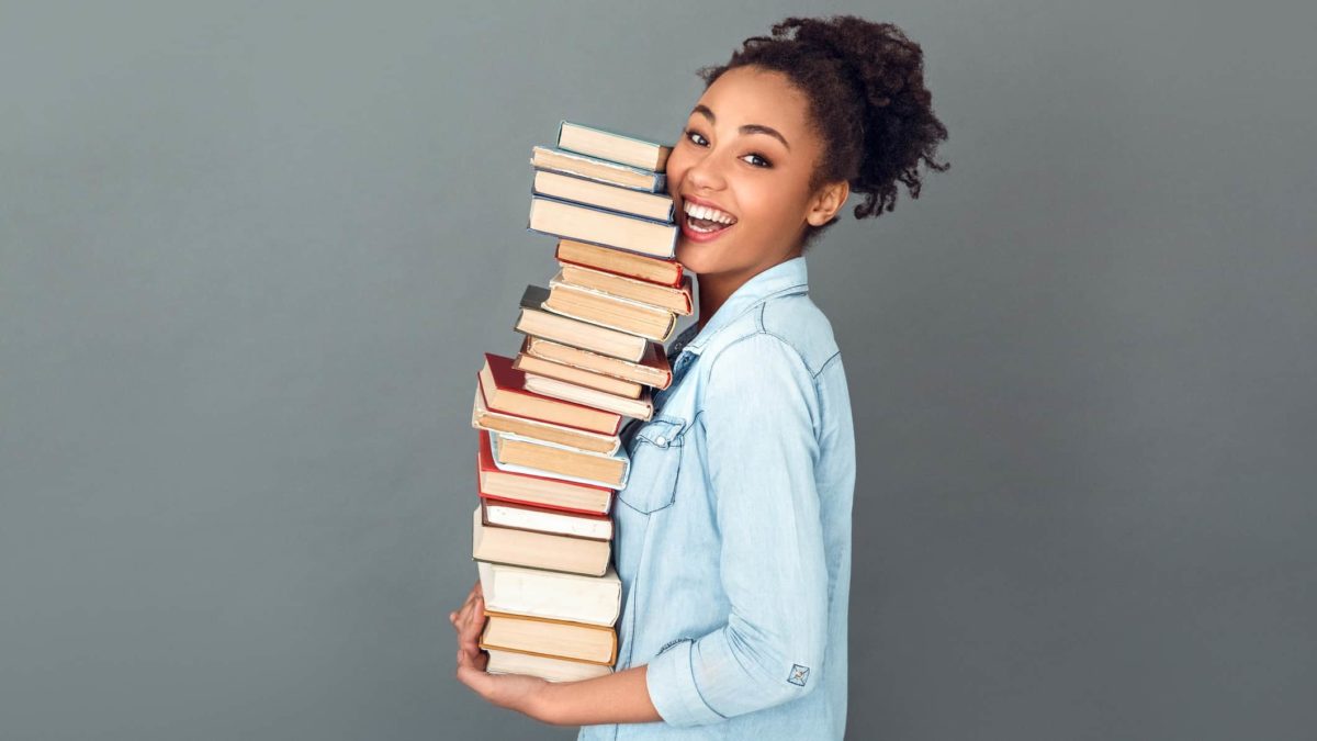 a smiling young woman carrying a pile of books, indicating a lifting share price for book sellers