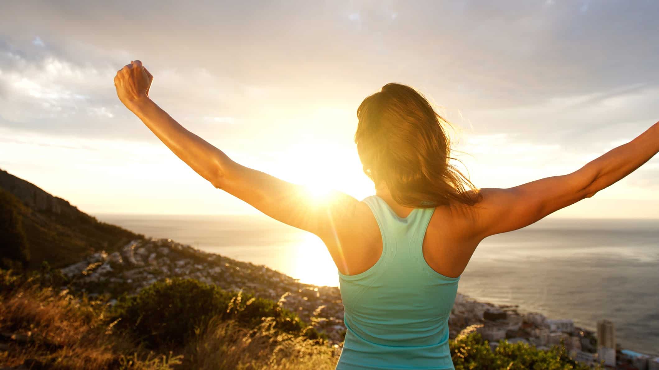 A fit woman stands on a hill facing the water at dawn with open arms embracing the future