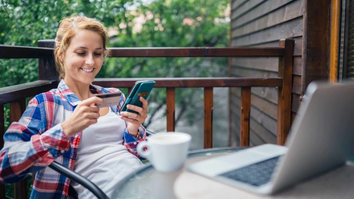 A happy woman sits on an outdoor deck with trees behind her and holds a credit card in one hand and her mobile phone in the other hand