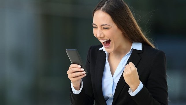 A happy woman looks at her mobile phone and fist pumps, indicating a share price rise