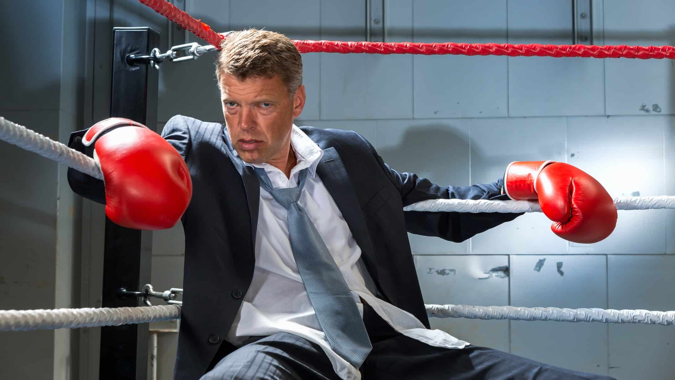 A businessman in a suit and wearing boxing gloves, slump in the corner of a ring, indicating a corporate fight between ASX companies