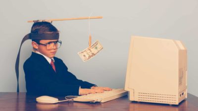 financial plans represented by boy sitting at old computer with dollar note hanging in front of his head