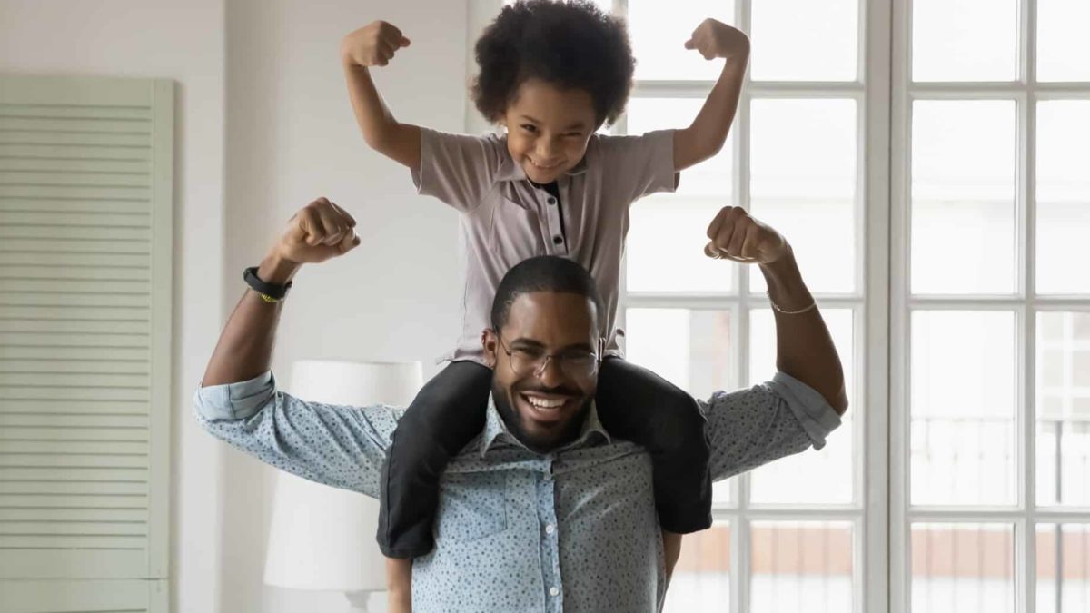 A young boy sits on his dad's shoulders while both flex their muscles.